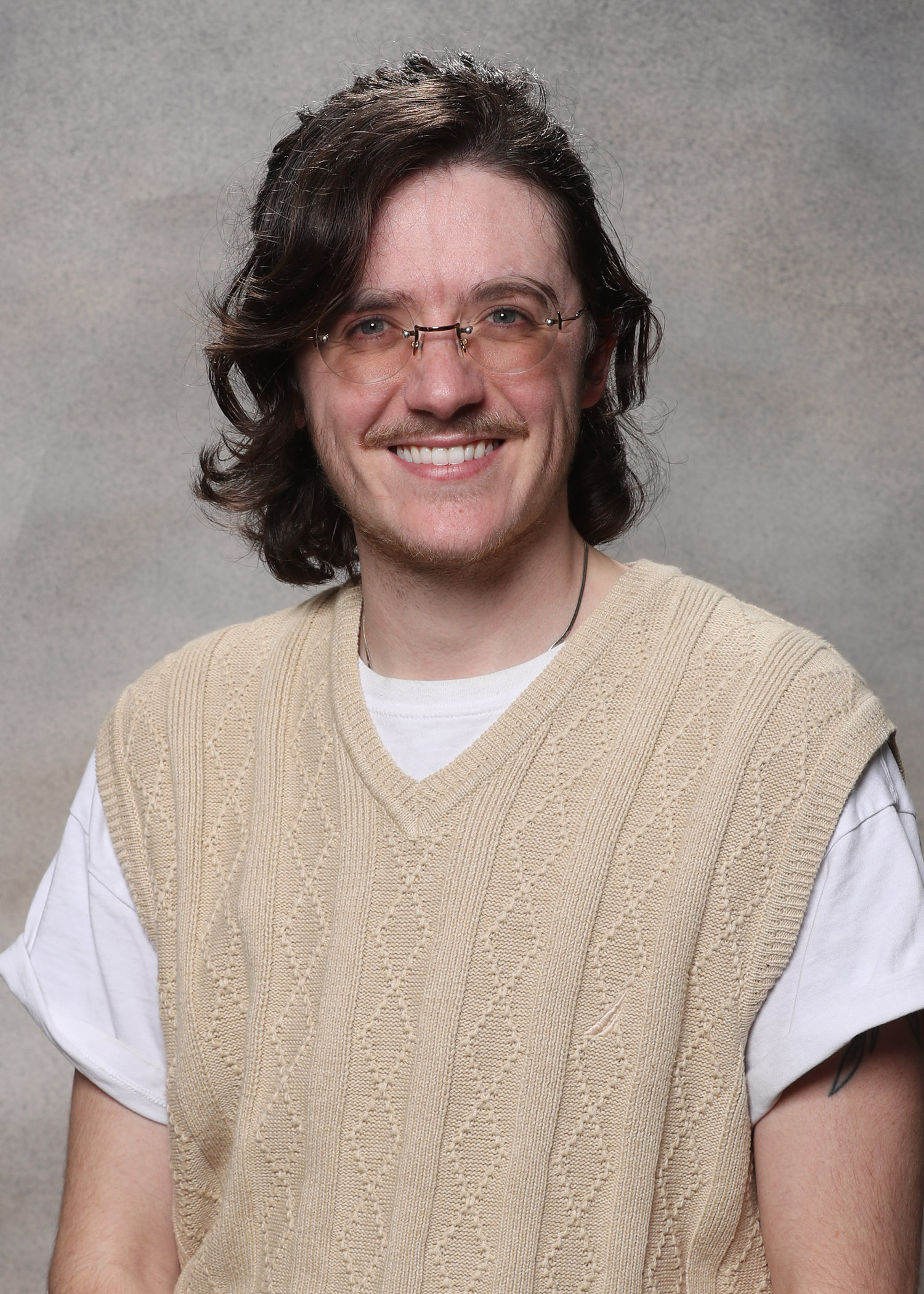 A young man wearing a beige crewneck sweater vest over a white t shirt with round glasses, smiling warmly.
